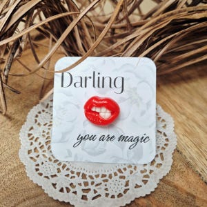 Könnte beinhalten: Eine rotlippenförmige Anstecknadel mit weißen Zähnen auf einer Karte mit der Aufschrift "Darling you are magic". Die Karte liegt auf einer weißen Spitzendecke, mit einem Holzgrund und Akzenten aus getrockneten Pflanzen.