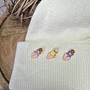 Op de afbeelding: Een crèmekleurige muts met drie kleine, kleurrijke spelden bevestigd. De spelden beelden figuren in badpakken af, met paarse, gele en blauwe ontwerpen. De muts is gemaakt van zacht, gebreid materiaal.