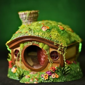 May include: A miniature hobbit-style house with a round front door, two small round windows, and a chimney. The roof is covered in green moss and small colorful flowers. The structure features brown wooden trim and is surrounded by floral landscaping.