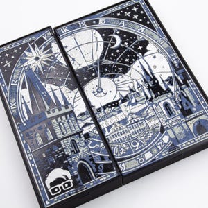 Op de afbeelding: Twee zwarte dozen met gedetailleerde blauw-witte illustraties van een stadsgezicht, hemelse elementen en een klok. Het ontwerp bevat architecturale details, sterren en een halve maan, met een gestileerde figuur in de hoek.
