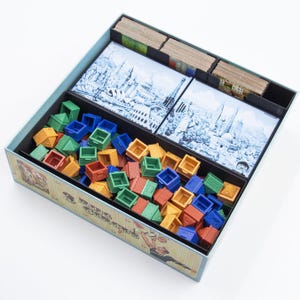 Könnte beinhalten: Eine Brettspielbox mit verschiedenen Komponenten. Die Box enthält bunte Miniaturhäuser in Blau, Grün, Orange und Rot. Es gibt auch illustrierte Karten und einen Boxteiler. Die Box hat eine hellblaue Außenseite und ein dekoratives Design an der Seite.
