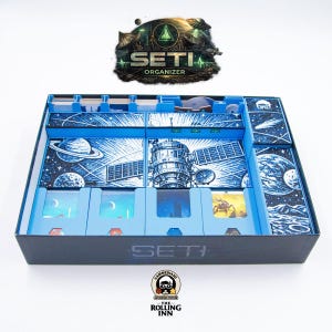 Pode incluir: Um organizador SETI azul e preto para um jogo de tabuleiro. O organizador apresenta um design detalhado com tema espacial com planetas, satélites e o texto "SETI ORGANIZER". A caixa é dividida em compartimentos.
