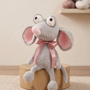 May include: A gray crocheted mouse plush toy with large, round eyes and pink-lined ears. It has a pink nose and a pink ribbon tied around its neck. The toy is sitting on a wooden block.