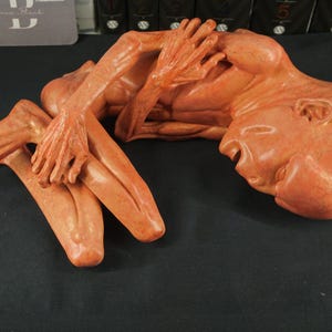 Op de afbeelding: Een terracotta-kleurige sculptuur die een menselijke figuur in een benarde pose uitbeeldt. De armen van de figuur zijn geheven, met de handen bij het hoofd en de mond open. De sculptuur staat op een donkere ondergrond.