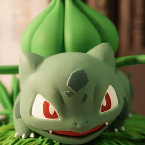 Może przedstawiać: Zielona i czerwona plastikowa figurka postaci Pokémona, Bulbasaura, z zielonym bulwą na plecach. Figurka siedzi na zielonej podstawie z detalami przypominającymi trawę.