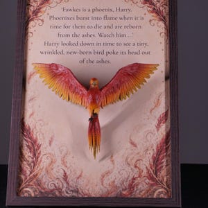 Può includere: Un'opera d'arte da parete incorniciata che presenta una fenice tridimensionale con ali rosse e arancioni centrata su uno sfondo in stile pergamena. Il testo recita: 'Fawkes is a phoenix, Harry. Phoenixes burst into flame when it is time for them to die and are reborn from the ashes. Watch him... Harry looked down in time to see a tiny, wrinkled, new-born bird poke its head out of the ashes.'