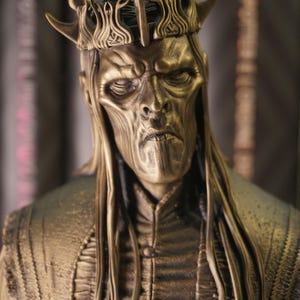 Może przedstawiać: Brązowe popiersie królewskiej postaci z koroną i długimi włosami. Szczegółowa rzeźba ma surowy wyraz twarzy i bogato zdobione szaty, sugerujące tematykę fantasy lub historyczną. Popiersie jest umieszczone na rozmytym tle.