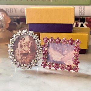 May include: Two small picture frames. The oval frame is bordered with clear, iridescent rhinestones and contains a sepia-toned image. The rectangular frame is bordered with pink floral-shaped rhinestones and contains a light-colored image.