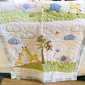 May include: A white and blue baby quilt with a jungle theme. The quilt features appliqued giraffes, elephants, monkeys, and clouds on a green and white background. The quilt has a blue and white striped border.