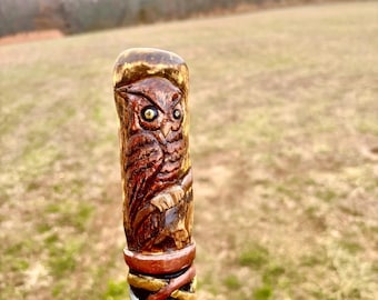 May include: A wooden walking stick with a carved owl design. The owl is brown with yellow eyes and is carved into the top of the stick. The stick has a series of decorative bands in brown, gold, and white.