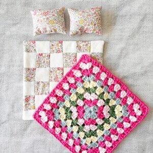 May include: A square crochet blanket featuring a vibrant pink border with a multi-coloured centre of pink, yellow, green and blue yarn, accompanied by a small patchwork quilt with a floral pattern and two matching miniature square pillows.