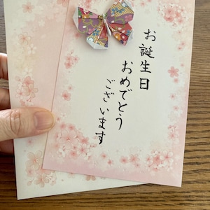 以下が含まれることがあります： ピンク色の誕生日カード。桜の模様と日本語の文字が書かれています。カラフルな折り紙の蝶がカードに付いています。カードは、お揃いの封筒の前に置かれています。