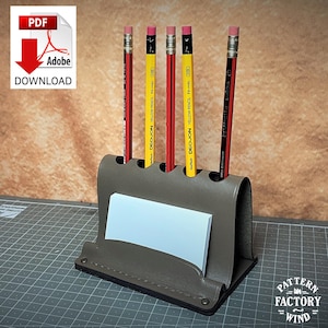 Puede incluir: Un organizador de escritorio de cuero gris con cinco lápices y un bloc de notas. Los lápices son rojos y amarillos con gomas de borrar rosas. El organizador tiene un diseño curvo y el texto "PDF Adobe DOWNLOAD".