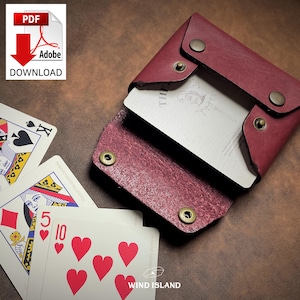 May include: A burgundy leather card holder with brass snap closures, holding a deck of cards. The cards feature a joker illustration and the text "THE". Playing cards with hearts and spades are visible, along with the text "WIND ISLAND".