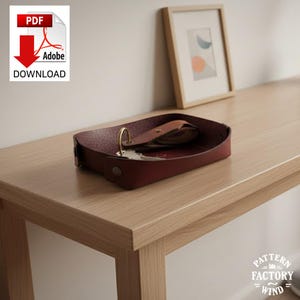 May include: A burgundy leather tray with a gold ring and keys, resting on a light wood surface. The tray is rectangular with rounded corners and a button closure. A framed abstract art piece is in the background. The image also includes a PDF download icon.