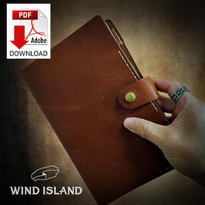 May include: A brown leather bound journal with a brass button closure and a leather strap. The journal is held in a hand with a ring on the finger. The text "WIND ISLAND" is visible at the bottom of the image.