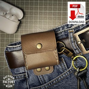 May include: A brown leather AirPods case attached to a belt loop on blue denim jeans. The case has a snap closure and is next to a key ring with keys. A white AirPods case is in the upper left corner. The image also includes a PDF download icon.