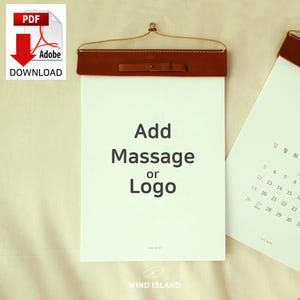May include: A brown leather hanging calendar with a white paper insert. The calendar has a hanging hook and a brown leather strap. The text on the calendar says "Add Massage or Logo".
