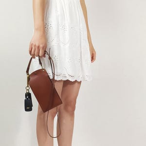 May include: A brown leather crossbody bag with a gold chain and a black key chain. The bag is worn by a person wearing a white eyelet dress.