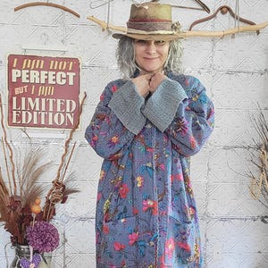 May include: A long-sleeved, quilted jacket in a muted blue hue featuring a vibrant floral pattern in shades of pink, yellow, and green. The jacket includes large front pockets, a button-down front, and wide, textured grey cuffs with visible stitching.