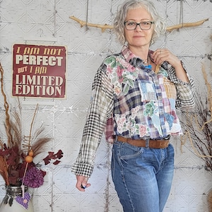 May include: A patchwork shirt with floral and plaid patterns, worn with blue jeans and a brown belt. The shirt has a mix of fabrics and textures. A sign on the wall reads "I AM NOT PERFECT BUT I AM LIMITED EDITION."