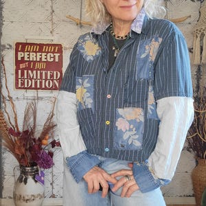 Può includere: Una camicia a righe blu e bianche con toppe floreali e un colletto in denim. La camicia ha un cartello che dice "Non sono perfetto, ma sono un'edizione limitata".