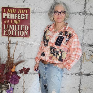 Puede incluir: Una mujer con una camisa patchwork con un diseño a cuadros naranja y blanco, combinada con vaqueros de mezclilla azul claro con parches y detalles desgastados. La camisa tiene parches de tela floral y de lunares. Un letrero en la pared dice "I AM NOT PERFECT BUT I AM LIMITED EDITION."