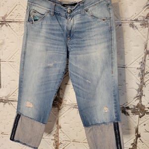 Peut inclure: Un jean en denim bleu clair avec des détails usés et une fermeture éclair noire sur les ourlets. Le jean a une coupe décontractée et une longueur raccourcie.