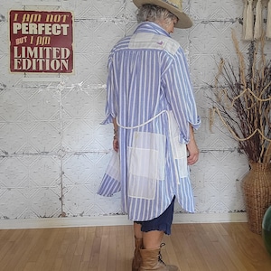 May include: A long, patchwork shirt with blue and white stripes and white squares. The shirt has a white belt and is paired with brown boots. A sign on the wall reads "I AM NOT PERFECT BUT I AM LIMITED EDITION."