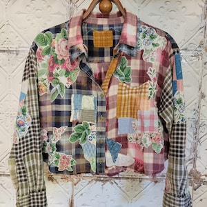 Könnte beinhalten: Ein Patchwork-Hemd mit Knopfleiste und einzigartigem Design. Das Hemd kombiniert florale, karierte und unifarbene Stoffflicken in Rosa, Blau und Grün. Es hat lange Ärmel und hängt an einem Holzbügel. Das Design ist eine Mischung aus Mustern.
