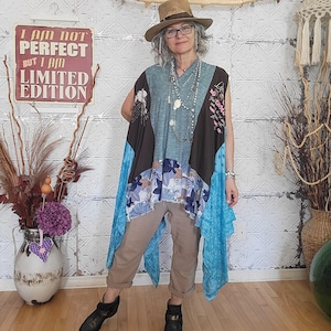 May include: A sleeveless tunic featuring a teal centre panel, brown side panels, and a blue floral patterned hem, paired with tan cropped trousers, a brown fedora hat, layered necklaces, and black ankle boots. The outfit is worn with glasses.