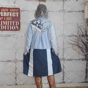 May include: A light blue denim jacket with a patterned back panel and a dark blue denim skirt with white stripes. The person is wearing brown leather boots and a grey cap. A sign reads "I AM NOT PERFECT BUT I AM LIMITED EDITION."