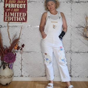 May include: White denim overalls with patchwork details on the knees. The overalls have a bib pocket and are paired with white sandals. A sign on the wall reads "I AM NOT PERFECT BUT I AM LIMITED EDITION."