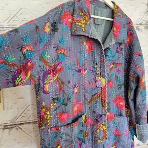 Peut inclure: Une veste matelassée grise ornée d'un motif floral et d'oiseaux aux tons vifs de rose, jaune et bleu sarcelle. Le vêtement présente un col, des manches longues à revers, deux poches avant et deux boutons violets sur le devant.