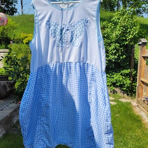 May include: A blue and white gingham dress with a butterfly motif. The sleeveless dress combines striped and checked patterns, with a light blue trim and a gathered waist. It is hanging on a white hanger outdoors, with a garden background.