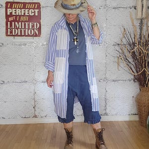 May include: A woman in a light blue and white striped long shirt, blue shorts, and brown boots. She is wearing a hat and necklaces. A sign on the wall says "I AM NOT PERFECT BUT I AM LIMITED EDITION."