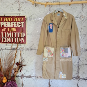 May include: A tan long-sleeve coat featuring a collar, a button-down front, and two large front pockets. The coat is decorated with various fabric patches, including floral and patterned squares, and lace trim accents on the pockets and hem.