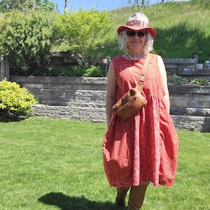 Può includere: Una persona indossa un vestito rosso fantasia, stivali marroni e un marsupio in pelle marrone. Indossa anche un cappello rosso con una fascia fantasia e occhiali da sole. Lo sfondo presenta erba verde, alberi e un muro di pietra.