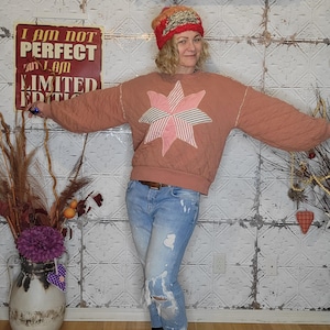 May include: A woman wearing a pink quilted sweatshirt with a large pink and white star applique, blue jeans with distressed details, and a red and orange knit hat. The sign behind her reads "I am not perfect but I am limited edition".
