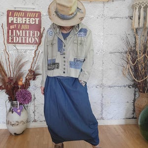 May include: A beige hooded jacket with blue bandana-print patchwork accents, a dark blue shirt, a long dark blue skirt, and a wide-brimmed tan hat. The jacket features a button-up front, chest pockets, and drawstrings. The outfit is paired with brown boots.