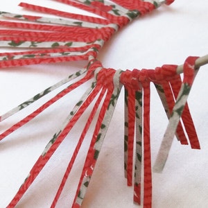 May include: A close-up of a red and white fabric fringe tied around a metal rod. The fringe is made of thin strips of fabric with a floral pattern.