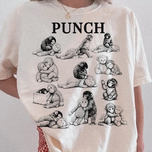 May include: Ivory-colored t-shirt with the word "PUNCH" in bold black letters. The shirt features a repeating pattern of black and white illustrations of monkeys in various poses. The shirt is made of a soft material.