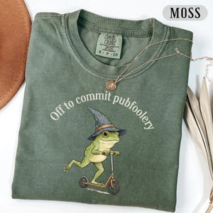 May include: Moss green t-shirt with the phrase "Off to commit pubfoolery" and a cartoon frog riding a scooter, wearing a wizard hat. A silver necklace with a pendant is draped across the shirt.