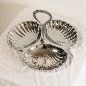 Puede incluir: Una bandeja plateada de tres secciones con asa. La bandeja tiene un diseño de concha y contiene joyas, incluyendo un anillo y una pulsera dorados. La bandeja está sobre una superficie blanca.