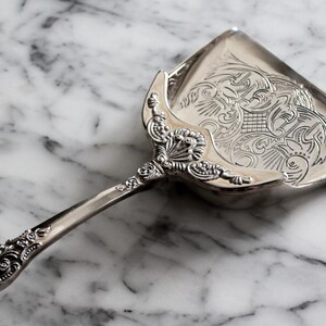 May include: A silver pie server with an ornate handle and a decorative, engraved blade. The blade features a detailed floral and scroll design. The server is resting on a marble surface.