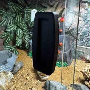 May include: A black rectangular handle with a recessed grip, possibly for a terrarium or aquarium door. The handle is mounted on a clear glass panel, with a background of artificial plants, rocks, and a decorative structure.