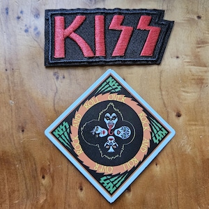 May include: Two patches featuring the band KISS. The top patch is rectangular with the word "KISS" in red block letters on a black background. The bottom patch is diamond-shaped with a band graphic and the words "KISS ROCK AND ROLL OVER".