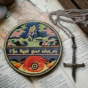 May include: A circular embroidered patch featuring a mountain landscape, a red dragon-like creature, and the text "The Road goes ever on" in yellow lettering, accompanied by a silver-toned sword pendant on a matching chain link necklace.