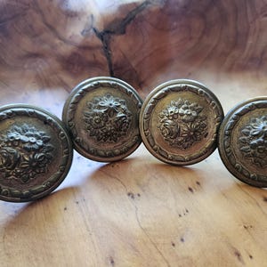 Peut inclure: Quatre boutons de meuble antiques de couleur bronze. Chaque bouton présente un motif floral détaillé dans un cadre circulaire. Les boutons sont disposés en rangée sur une surface en bois, mettant en valeur leur esthétique vintage.