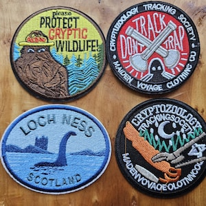 Könnte beinhalten: Vier gestickte Aufnäher mit Designs zum Thema Kryptiden. Einer trägt die Aufschrift "Protect Cryptic Wildlife!" mit einer Bigfoot-Illustration. Ein anderer sagt "Track Don't Trap" mit gekreuzten Schaufeln. Ein dritter zeigt das Ungeheuer von Loch Ness.